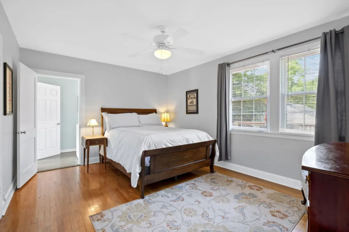 A peaceful bedroom scene featuring a neatly made bed with soft linens, a nightstand with a glowing lamp, a window dressed with curtains letting in natural light, and a cozy rug underfoot. The ambiance is serene and inviting, ideal for relaxation and restful sleep.