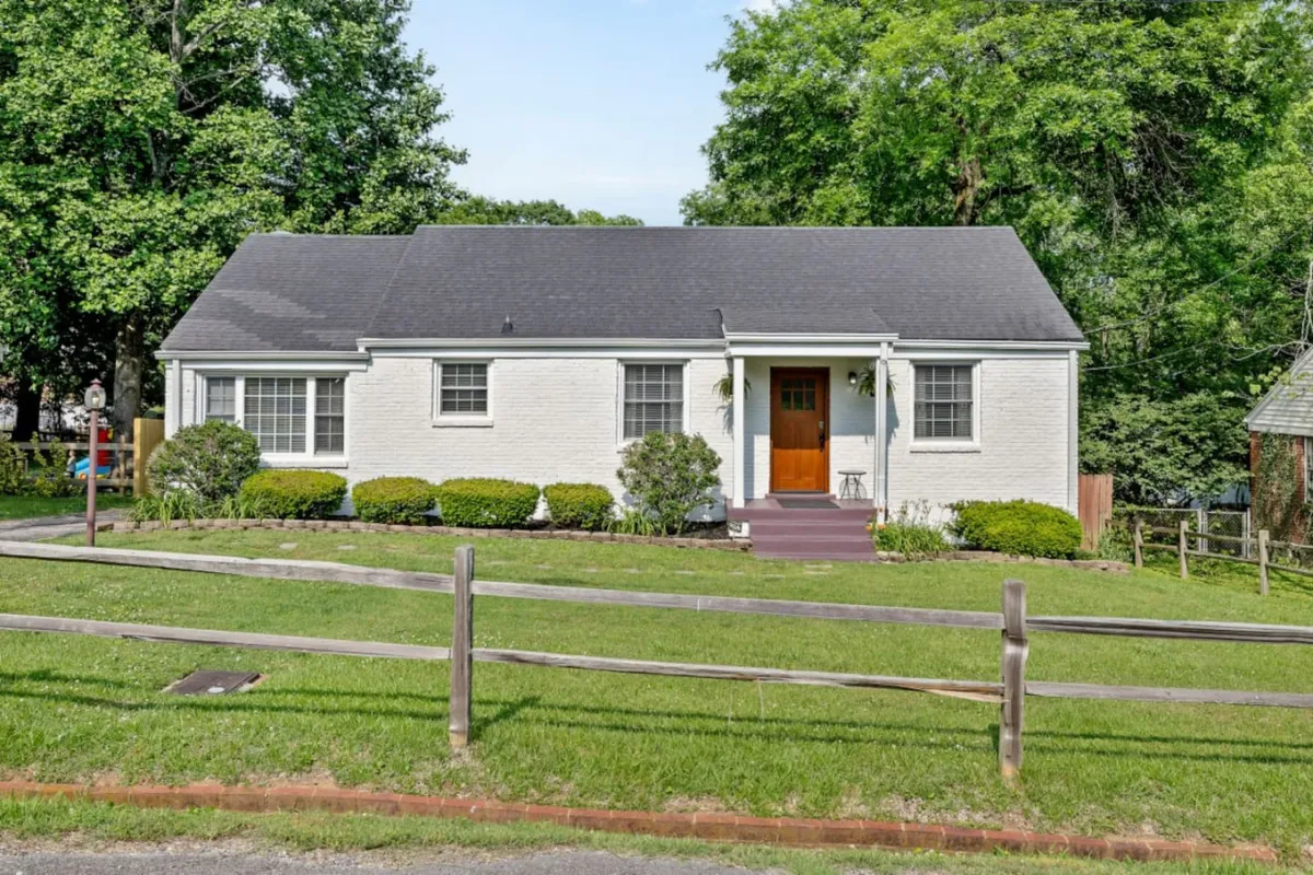 The image shows the inviting exterior of a single-family house in East Nashville, featuring a welcoming front door, a beautifully landscaped garden, and a spacious, fenced yard perfect for outdoor activities. The scene is filled with warm, natural light, suggesting a serene and family-friendly environment ideal for relaxation and gatherings.