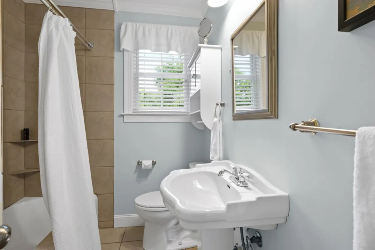A fresh, tranquil bathroom with a modern sink and elegant faucet, a white-curtained shower area, a framed mirror, and a shelf neatly stocked with towels. The atmosphere promotes relaxation and rejuvenation, supporting the morning or evening routines of guests.