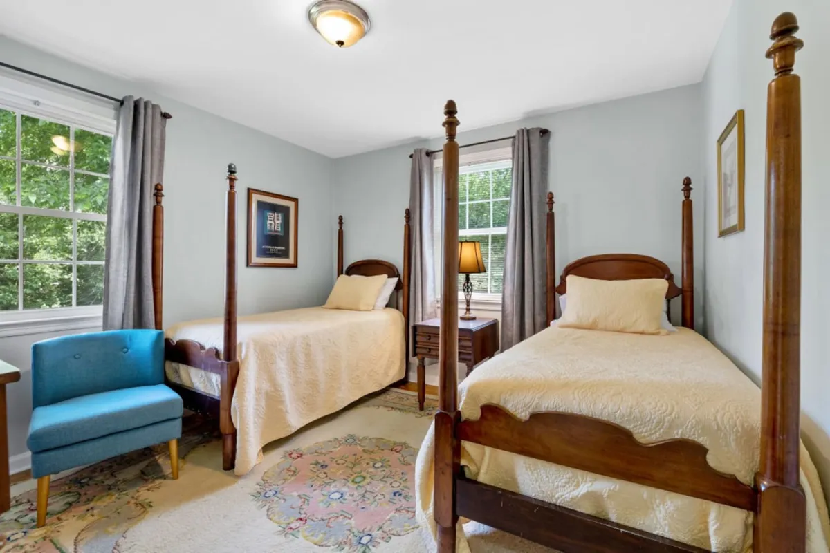 A calm, cozy bedroom with two wooden twin beds, a blue accent chair, a reading lamp on a side table, and a decorative rug. The space is inviting for both rest and quiet conversation, with a design that suits younger guests or friends.