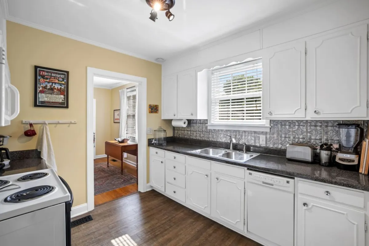A warmly lit kitchen featuring white cabinetry, sleek black granite countertops, and modern appliances. Natural light streams in through a window, illuminating an inviting workspace ideal for meal prep and gatherings. Decorative wall art adds a touch of personality, and the counters are spacious enough for shared cooking activities.